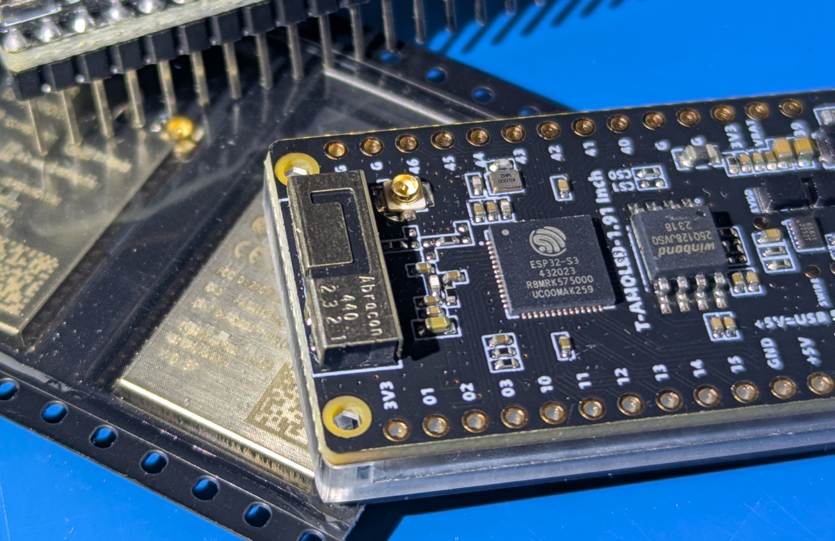 Close-up of a black ESP32-S3 development board on a blue mat, showing the square ESP32-S3 SoC beside a Winbond SPI flash chip, a U.FL antenna connector and gold castellated pin headers.