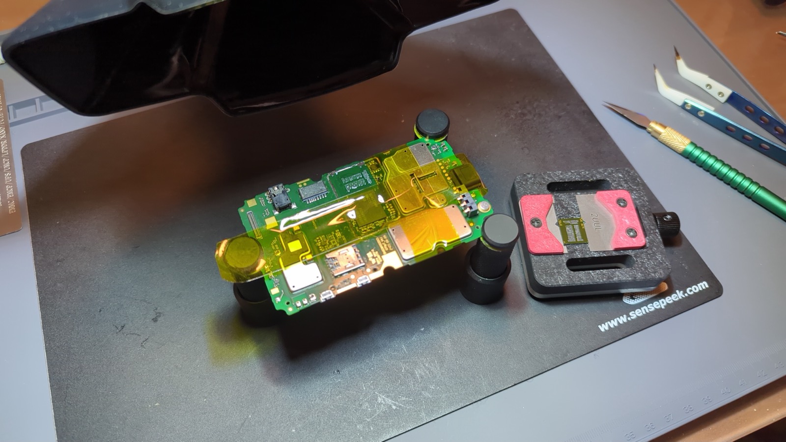 Phone mainboard masked with kapton tape under a hot-air nozzle, with the removed eMMC chip sitting in a holder beside it