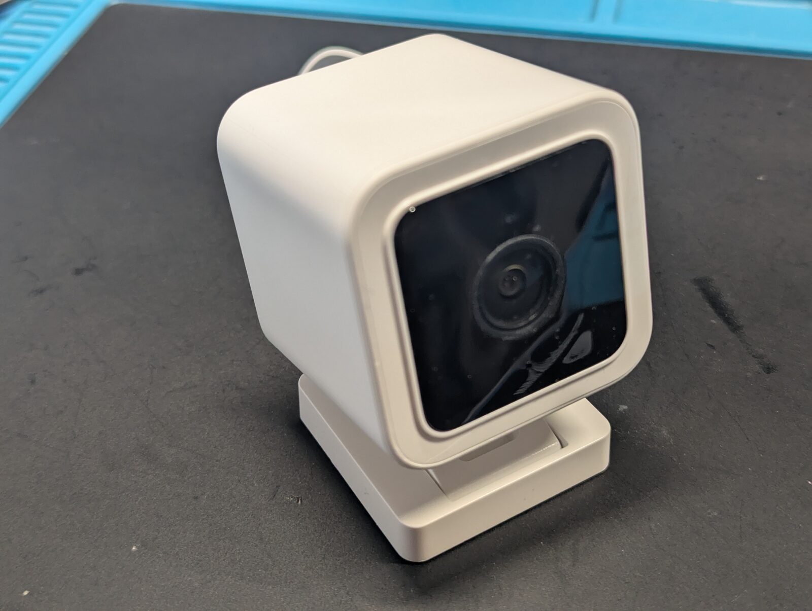 A white Wyze Cam v3 cube camera on its stand, sitting on a dark workbench mat.